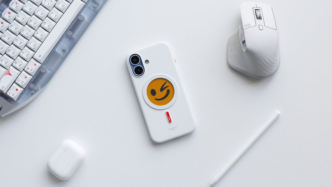 White XTREM instant custom phone case featuring a playful winking emoji design on a clean desk setup with keyboard, mouse, stylus, and earbuds.